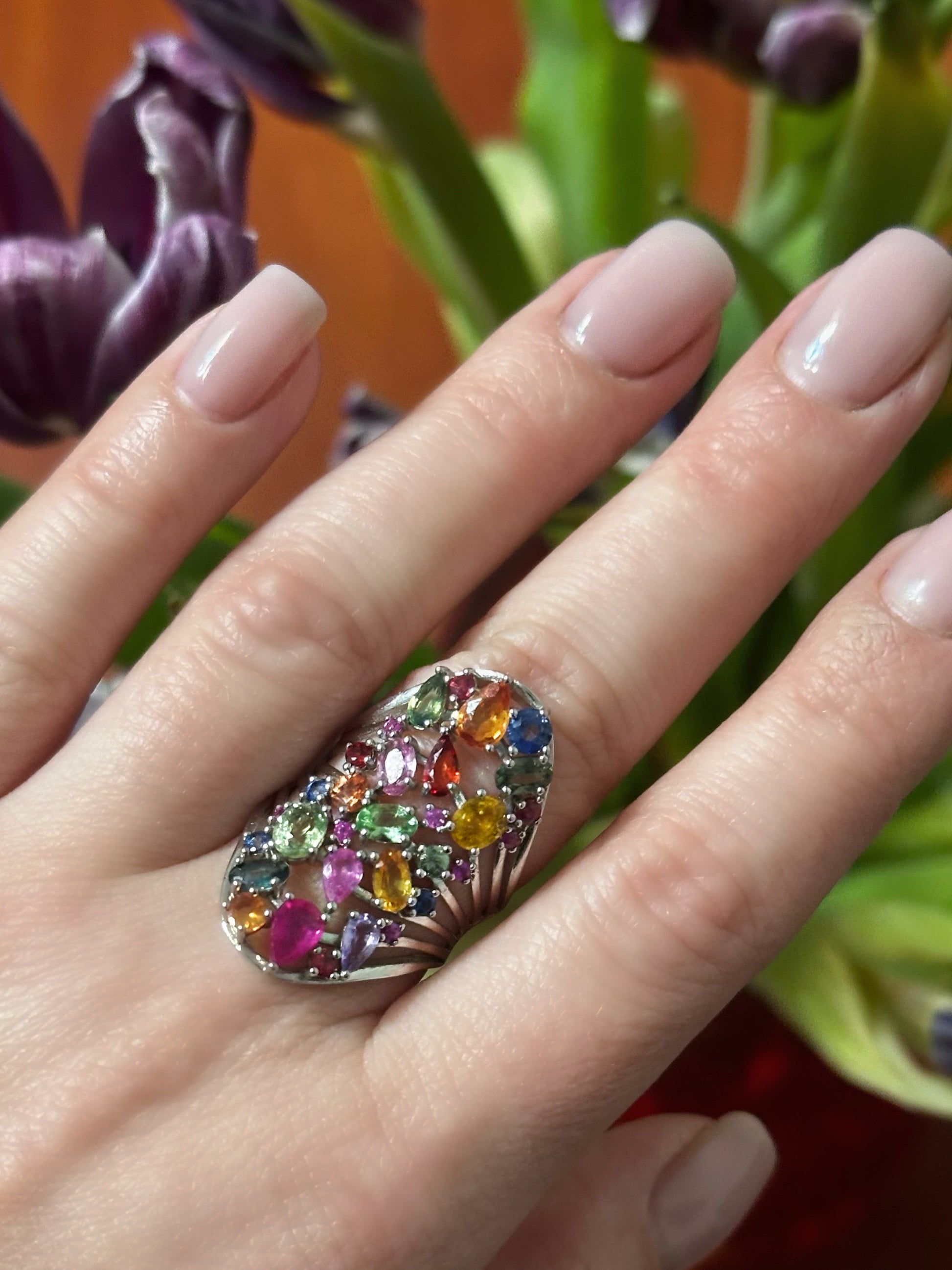 Multicolored gemstone ring on a hand with flowers in the background